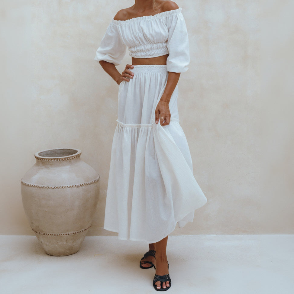 Woman wearing a white off-shoulder top and skirt standing against a plain background.
